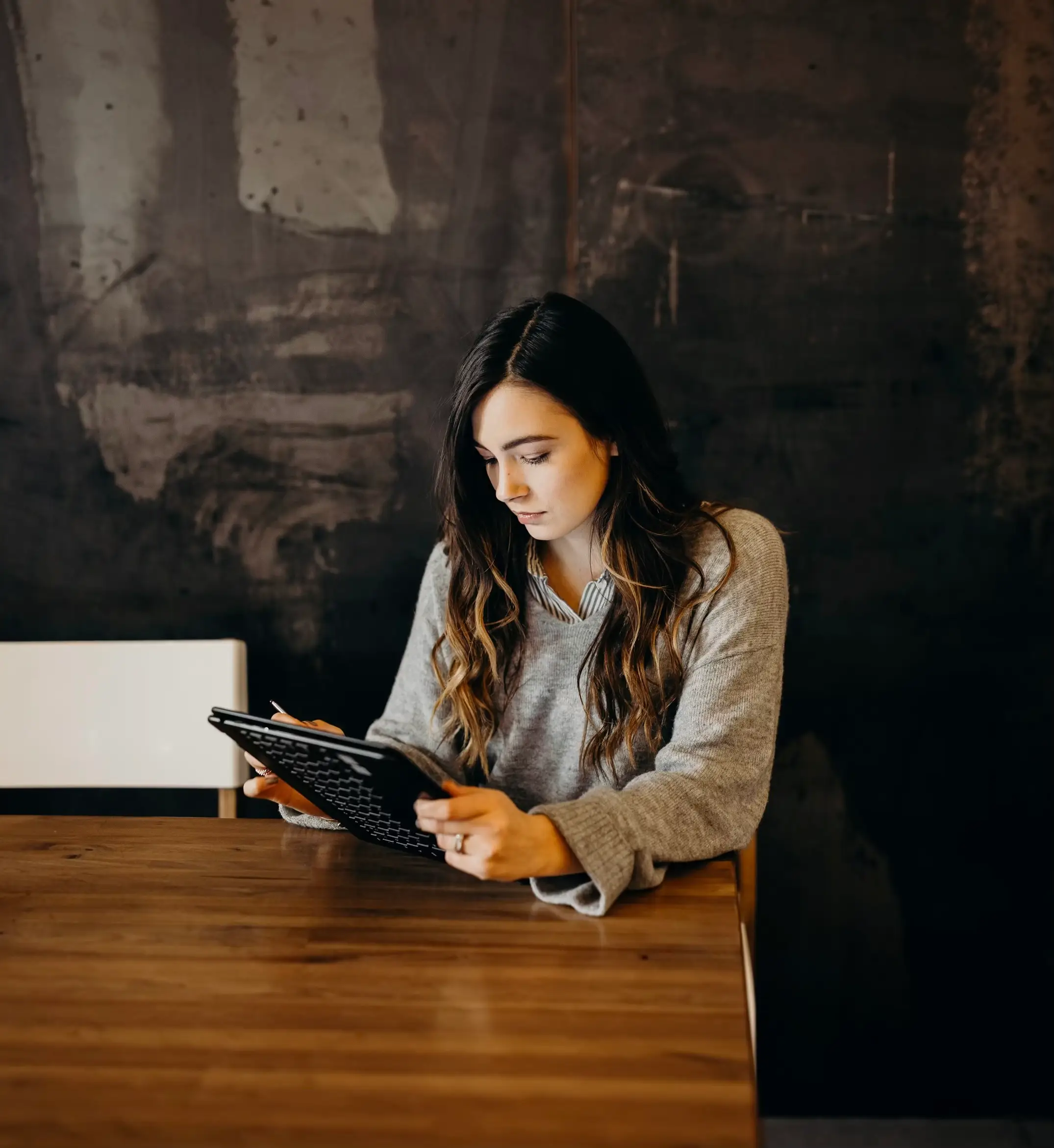 A woman at a wooden table looks down at a tablet in her hands, in front of a dark weathered wall.